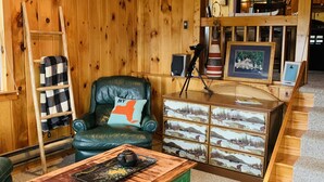 A cozy room with wooden walls, a green armchair with a pillow, a wooden coffee table, a ladder, a dresser with drawers, and a staircase. - Waterfront Camp with Boathouse - Wifi, kayaks, canoe and sandy bottom out front. (Old Forge)