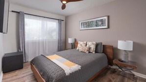 Une chambre à coucher moderne, avec un lit, des lampes de chevet, un ventilateur de plafond, une fenêtre avec des rideaux et une œuvre d’art encadrée accrochée au mur.