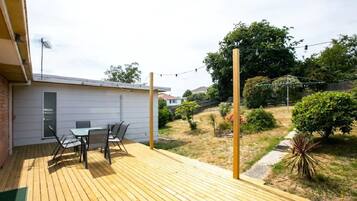 A wooden deck with a table and chairs, a small building with a door, and a garden with trees and shrubs.