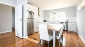 A modern kitchen with white cabinets, a stainless steel refrigerator, and a dining area with a wooden table and white chairs. - Family Escape | Views & Wi-Fi (Newstead)