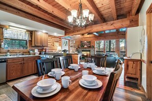 A wooden cabin interior with a dining table set for four, wooden cabinets, a chandelier, and a view of the outdoors.