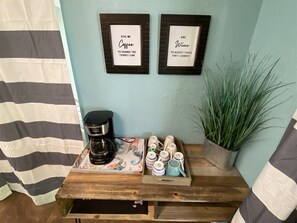 A coffee station with a coffee maker, mugs, and a tray of coffee cups. There are two framed quotes on the wall.