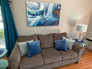 A living room with a grey sofa, two blue cushions, a teal curtain, a wall-mounted abstract painting, and a small round table with a lamp. - 4 Bedroom Lakeview home next to Margaritaville (Osage Beach)