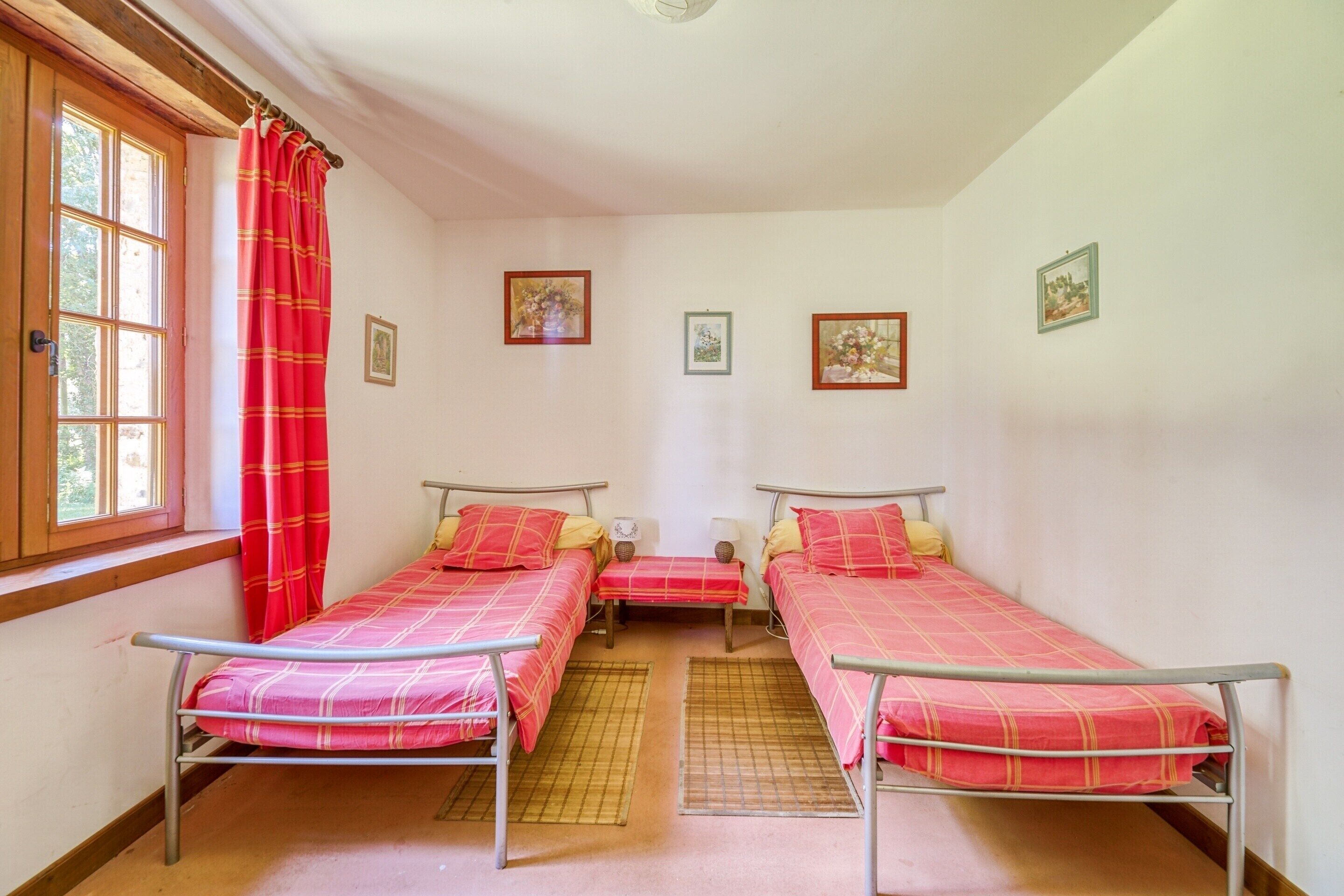 A room with three metal beds, a window with red curtains, and several framed pictures on the walls.