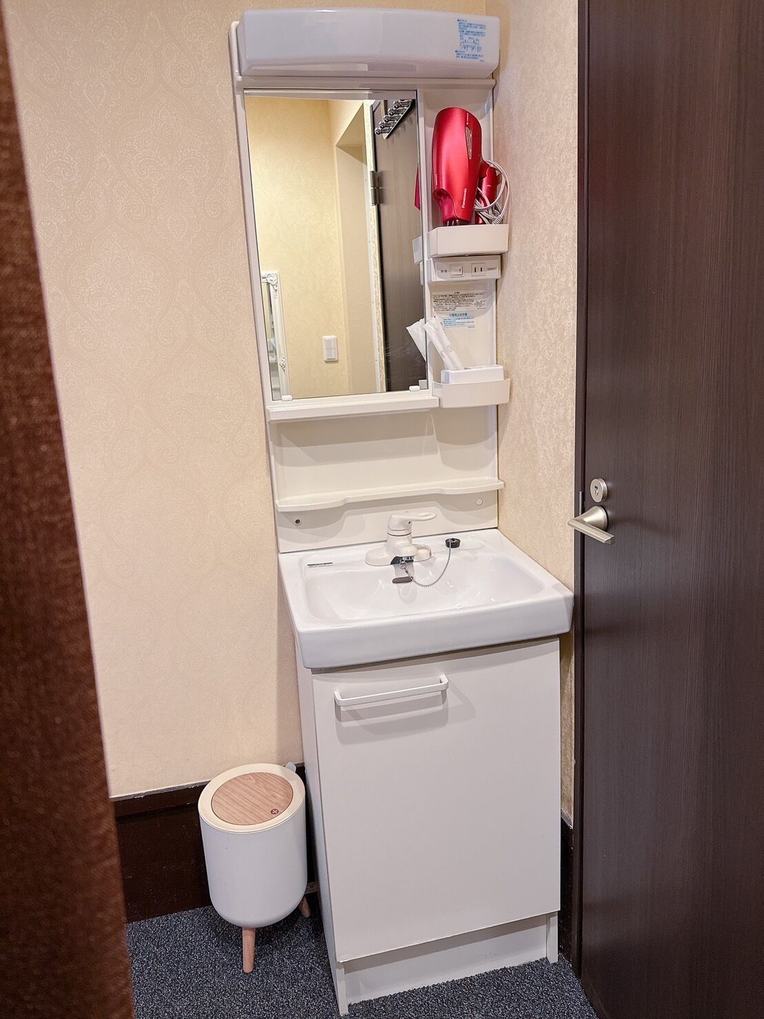 Un baño pequeño con un lavamanos blanco, un espejo y un mueble de almacenamiento. Se ve un secador de pelo rojo en el reflejo del espejo.