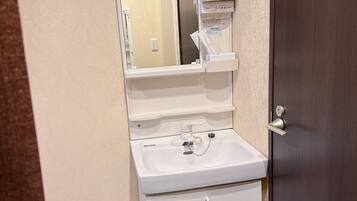 Un baño pequeño con un lavamanos blanco, un espejo y un mueble de almacenamiento. Se ve un secador de pelo rojo en el reflejo del espejo.