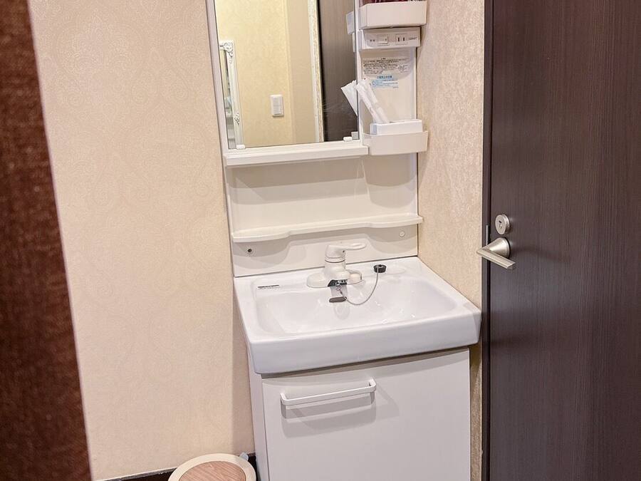 Un baño pequeño con un lavamanos blanco, un espejo y un mueble de almacenamiento. Se ve un secador de pelo rojo en el reflejo del espejo.