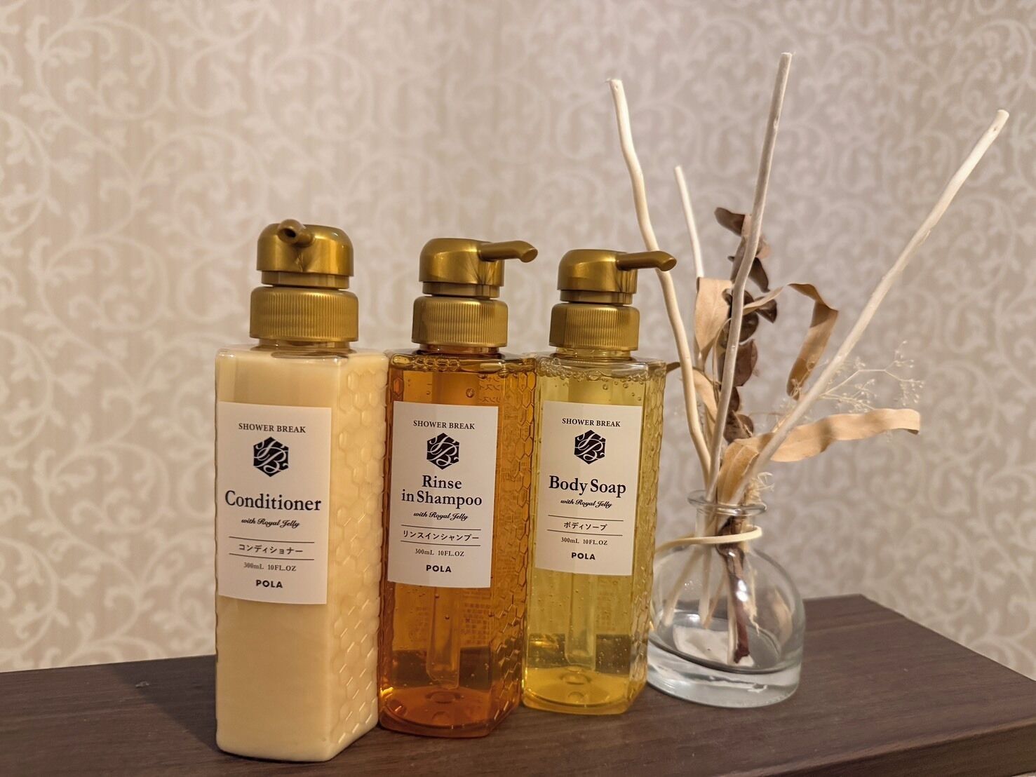 Three bottles of shower products on a wooden surface with a patterned wallpaper background.