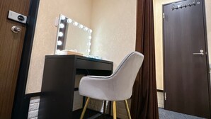 Habitación de esquina con escritorio, silla, tocador con luces y una puerta.
