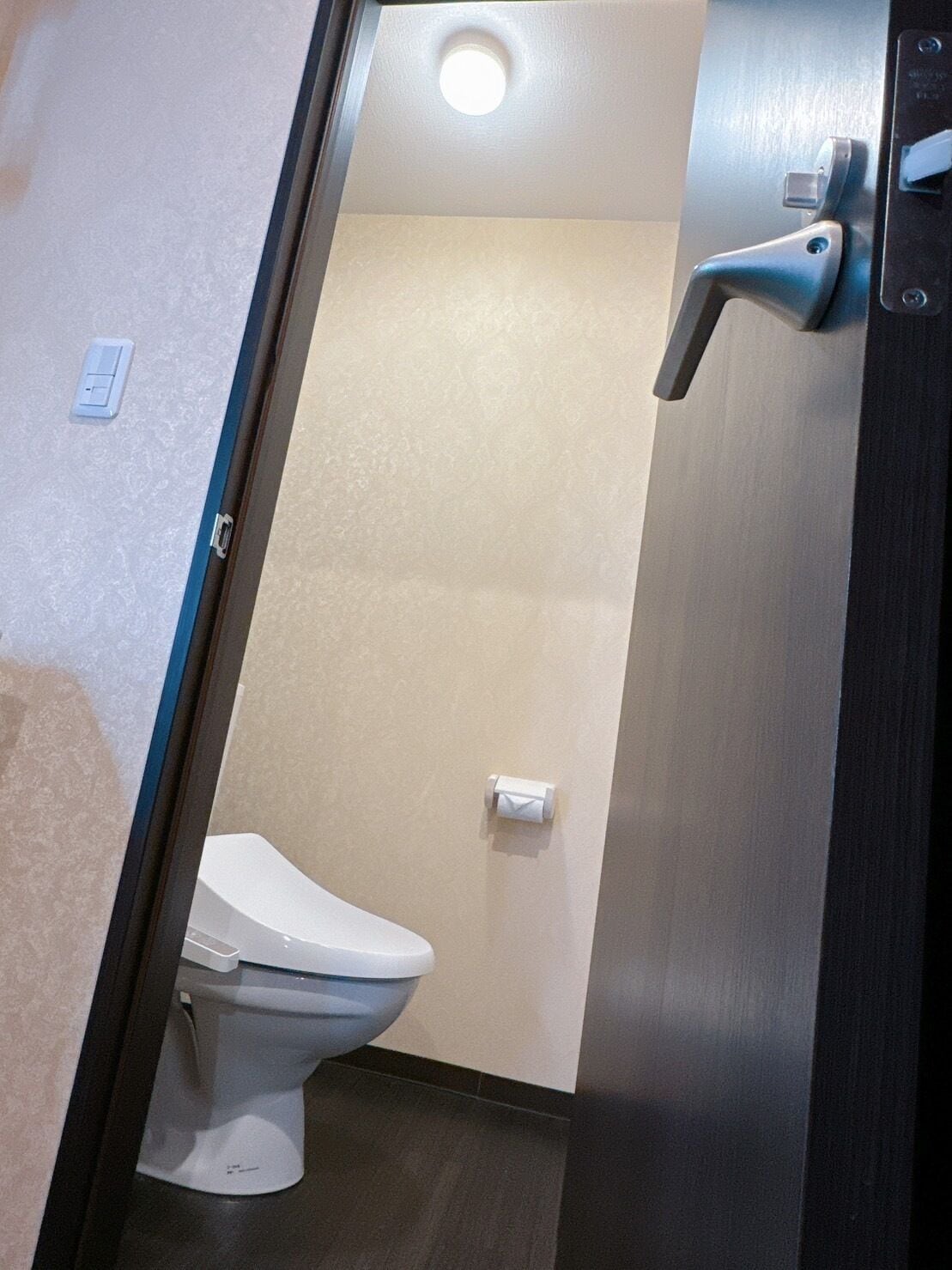 A small, narrow bathroom with a toilet, a towel dispenser, and a light fixture on the ceiling.