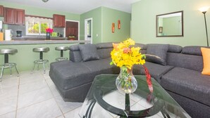 Ein modernes Wohnzimmer mit einem grauen Sofa, einem Glastisch und einer Vase mit Blumen. Im Hintergrund befinden sich zwei Barhocker und eine Küche.