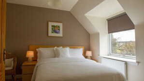 Un dormitorio con techo inclinado, un ventanal con vistas, una cama con ropa blanca, dos lámparas de noche y un cuadro en la pared.