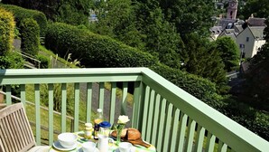 Una mesa con vajilla blanca, un mantel a cuadros verde y amarillo, y una botella en un balcón con barandilla verde, con vistas a un pueblo.