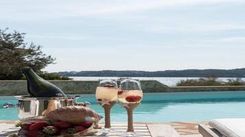 A poolside setting with two glasses of a beverage, a bottle, and a bowl of strawberries.
