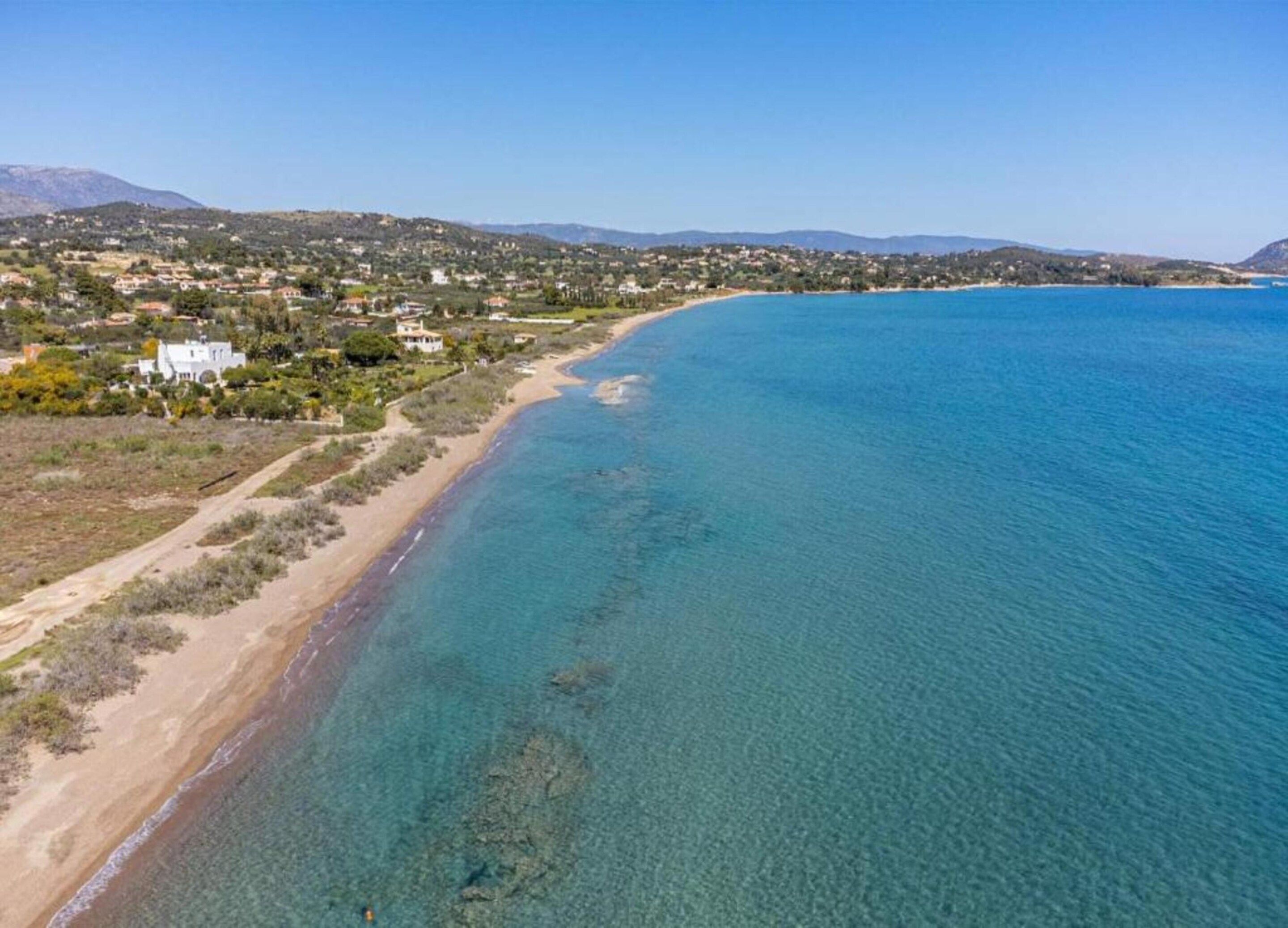 A coastal landscape with a clear beach, a mountain range in the background, and a residential area with houses.
