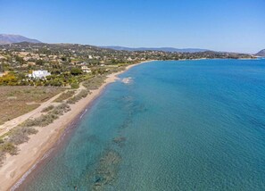 A coastal landscape with a clear beach, a mountain range in the background, and a residential area with houses.
