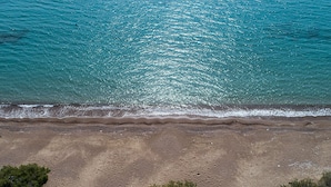 An aerial view of a beach with clear turquoise water, sandy shore, and sparse vegetation.