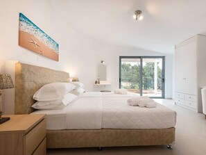 A modern bedroom with a large bed, a bedside table, a lamp, a painting of a beach scene, a mirror, and a sliding glass door leading to an outdoor area. - Villa in Peloponnese With Pool (Ermionida)