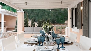 An outdoor dining area with white furniture, blue glassware, and a view of a mountainous landscape. - Villa in Peloponnese With Pool (Ermionida)