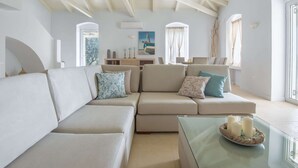 A spacious living room with a high ceiling and exposed beams, a large beige sectional sofa, a glass coffee table with candles, and a dining area in the background. - Villa in Peloponnese With Pool (Ermionida)