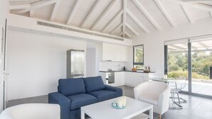 A modern living room with a blue sofa, white armchairs, and a white coffee table. A kitchen with white cabinets and a window is visible in the background. - Holiday Home in Porto Heli for 6 Guests (Ermionida)