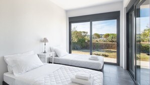 A modern bedroom with two beds, a nightstand, a lamp, and a large sliding glass door leading to a garden. - Holiday Home in Porto Heli for 6 Guests (Ermionida)