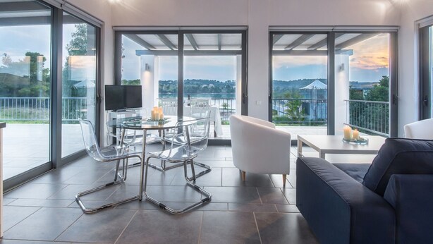 A modern living room with a glass table, white chairs, a blue sofa, and large sliding glass doors offering a view of the outdoors. - Holiday Home in Porto Heli for 6 Guests (Ermionida)