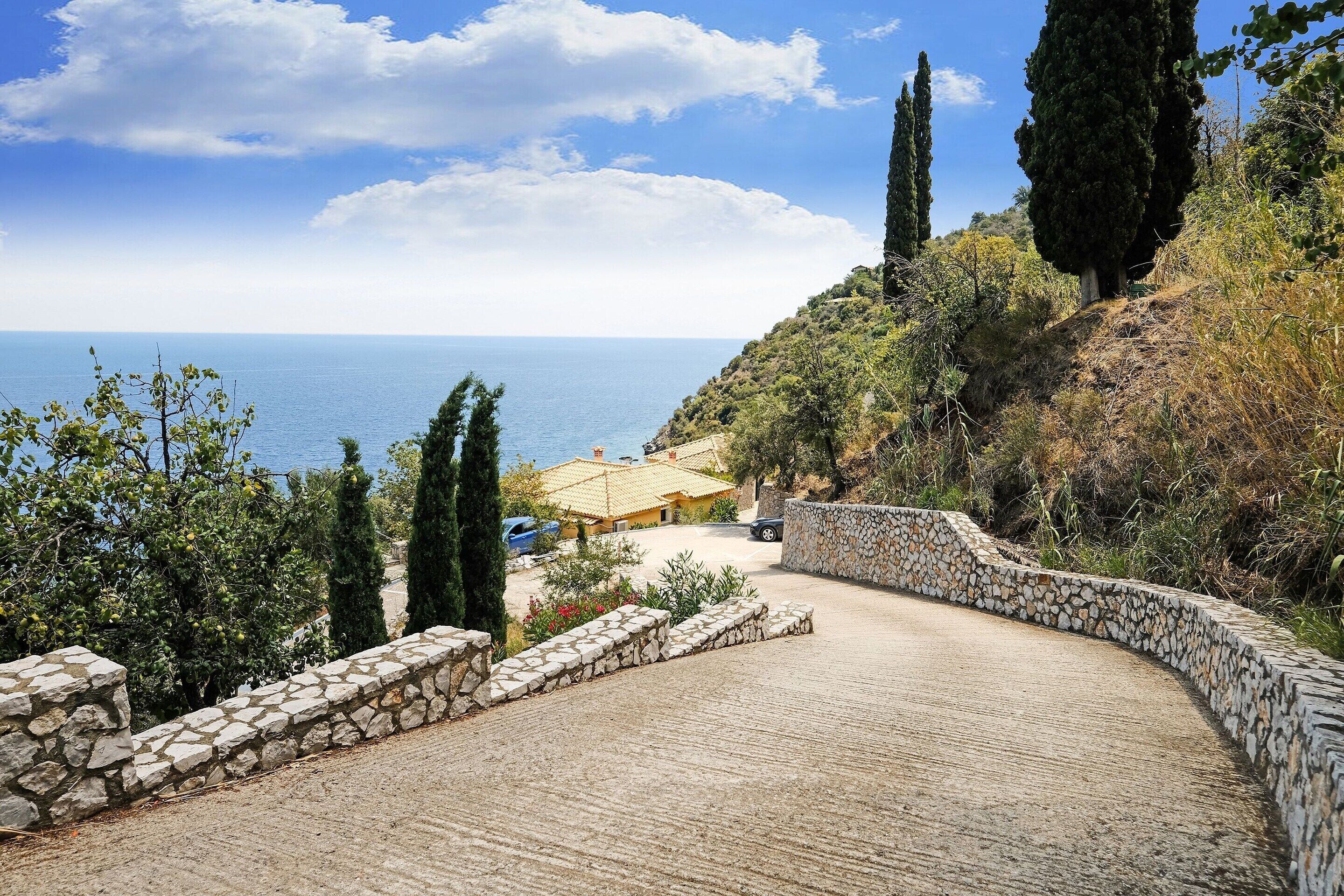 Un sentier pavé mène à une zone côtière bordée d’un mur de pierres et de végétation.