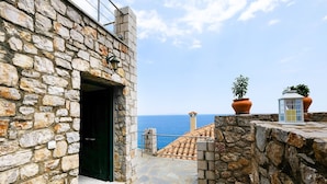 Un bâtiment en pierre offrant une vue sur la mer, un chemin bordé de carreaux et des plantes en pot.