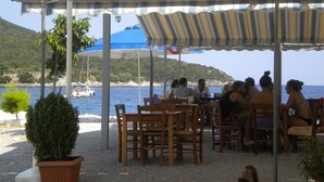 An outdoor seating area with tables and chairs under striped awnings, overlooking a body of water with boats and a hilly landscape in the background. - Chalet in Leonidio Near Mount Parnon (South Kynouria)