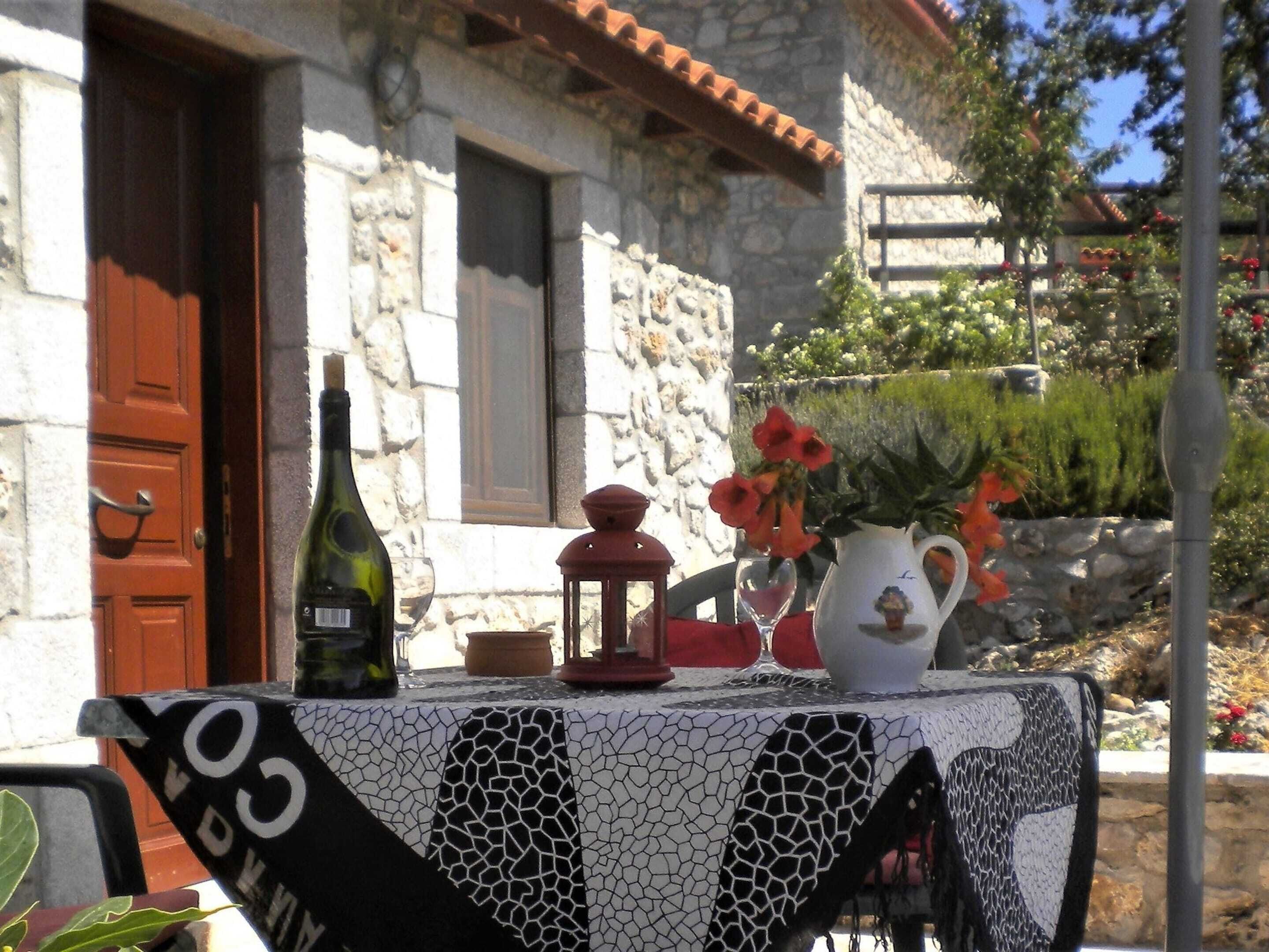 A table with a bottle of wine, a lantern, and a vase with flowers outside a stone building.