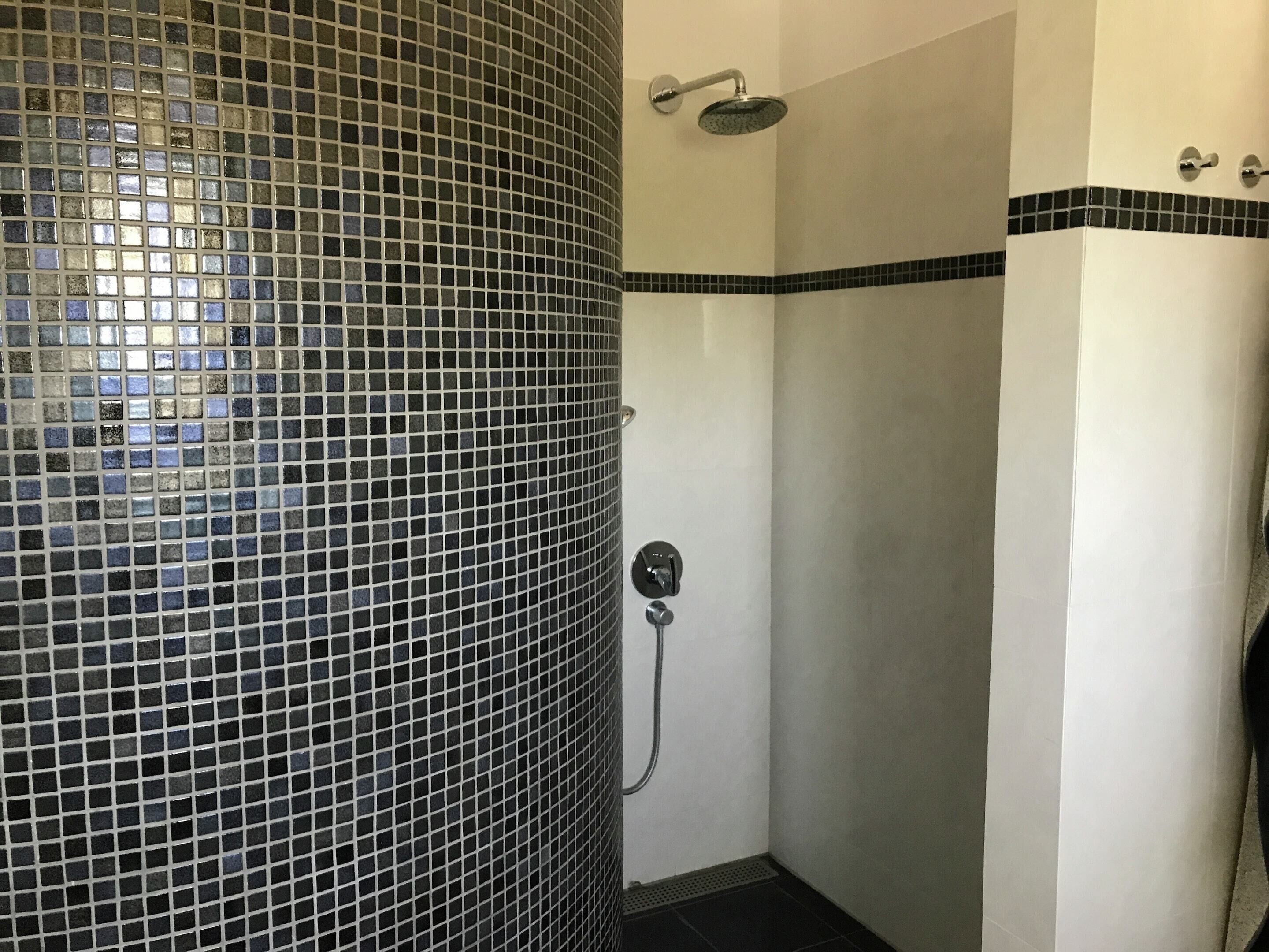 A bathroom with a tiled shower wall, a handheld showerhead, and a wall-mounted faucet.