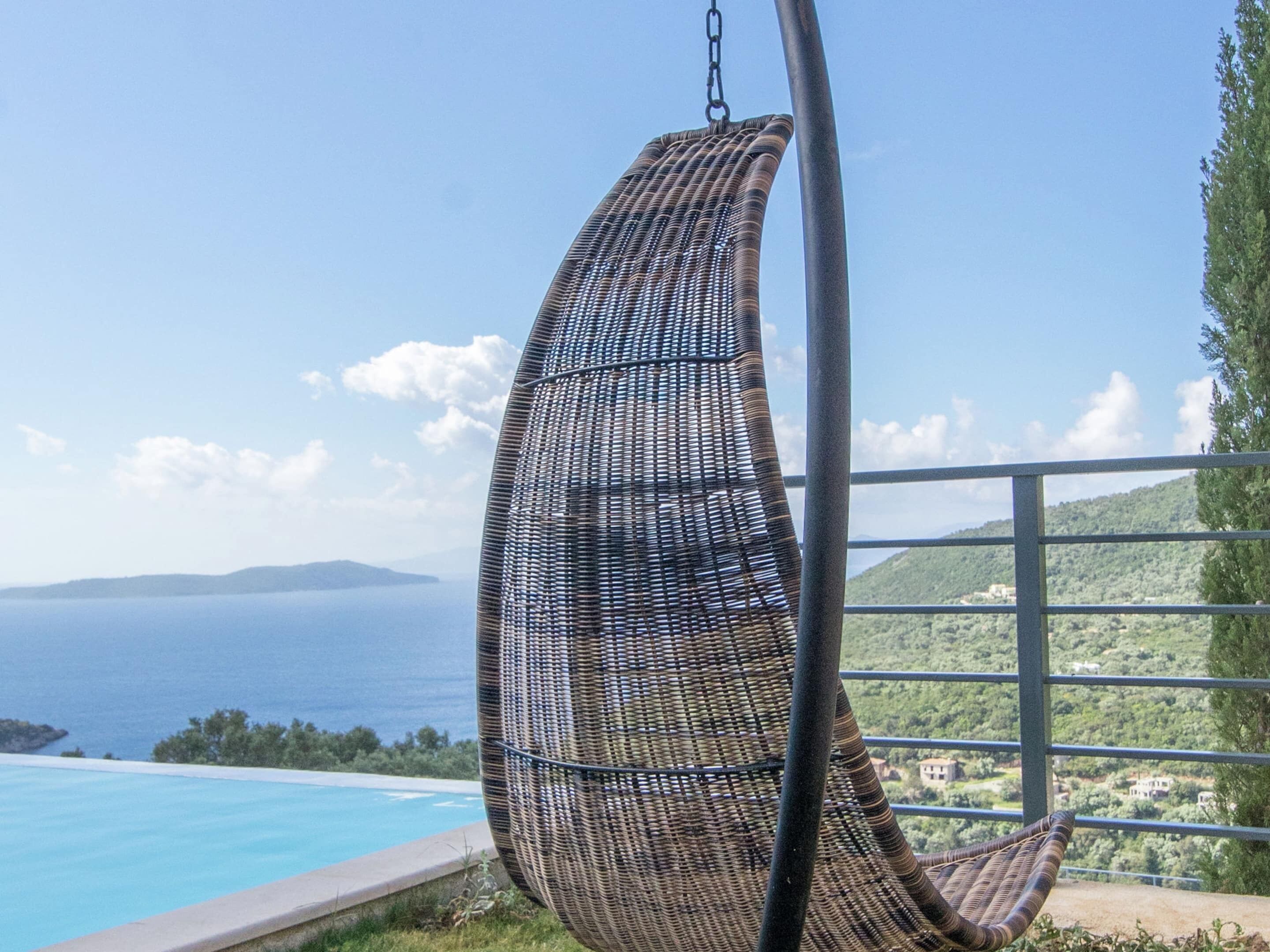 A wicker hanging chair overlooking a pool and a scenic view of the sea and hills.