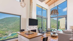 A spacious living area with a large TV, wooden flooring, and a view of a mountainous landscape through large windows. - Villa in Sivota With Private Pool & Beaches (Lefkada)