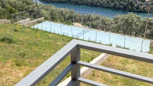 A tennis court with a metal railing in the foreground, surrounded by greenery and a view of a coastal area with houses. - Villa in Sivota With Private Pool & Beaches (Lefkada)
