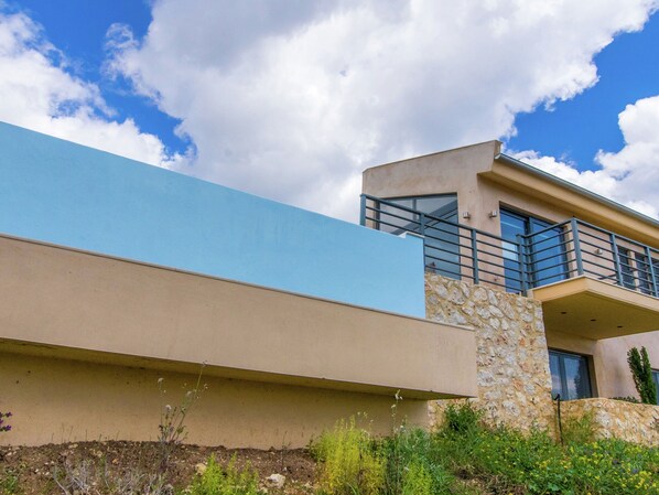 A modern building with a blue facade, a stone base, and a balcony with a metal railing. - Villa in Sivota With Sea View & Pool (Lefkada)