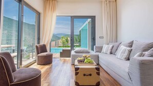 Un soggiorno moderno con divano, poltrona e poggiapiedi, un tavolino in legno con frutta, e vista su una piscina e montagne attraverso ampie finestre.
