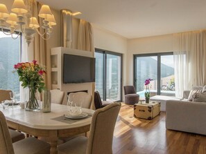 Habitación de hotel moderna con zona de comedor, sofá, televisor y vistas a las montañas.