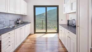 Una cucina moderna con mobili bianchi, un piano di lavoro nero e un pavimento in legno. È presente un'ampia porta vetrata scorrevole che dà su un balcone con vista su un pendio verde.