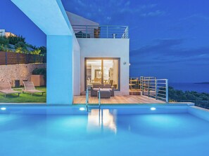 Un espace moderne aménagé au bord de la piscine, avec un salon clos par des baies vitrées, des sièges d’extérieur et une vue sur la mer.