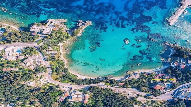 Luftaufnahme einer Küstenlandschaft mit klarem türkisblauem Wasser, einem Sandstrand und vereinzelten Gebäuden.