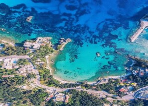 An aerial view of a coastal area with clear turquoise waters, a sandy beach, and scattered buildings. - Tranquil Apartment in Paleokastrites with Pool & Garden near Sea (Corfu)