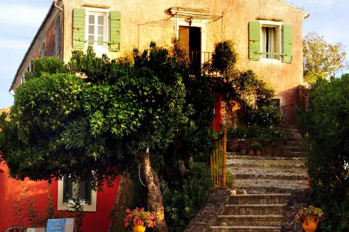 A rustic building with a staircase leading to a door, surrounded by lush greenery and a red wall.