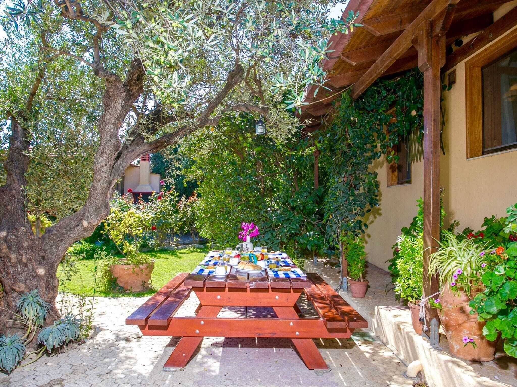 An outdoor dining area with a wooden table set for two, surrounded by lush greenery and potted plants.