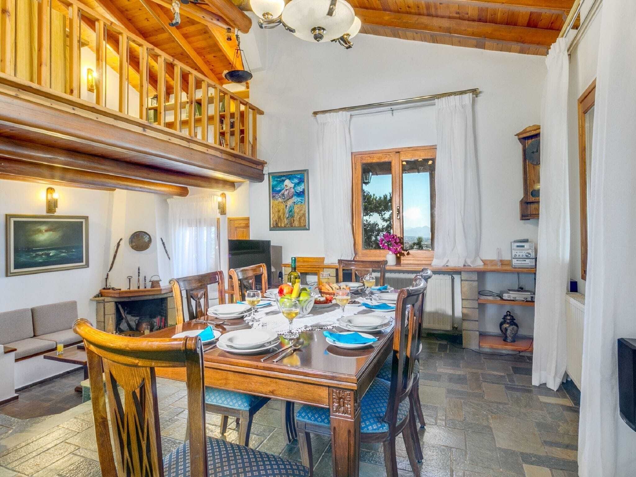 A dining area with a wooden table set for four, surrounded by chairs. A fireplace is visible in the background, and there is a window with a view of the outdoors.