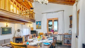 Uma sala de jantar com uma mesa de madeira posta para quatro pessoas, cercada por cadeiras. Uma lareira é visível ao fundo, e há uma janela com vista para o exterior.