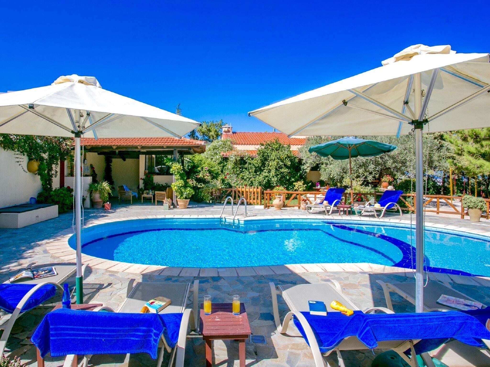 A pool area with lounge chairs and umbrellas, surrounded by greenery and a building with a tiled roof.