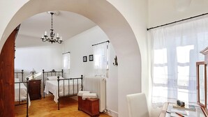 A bedroom with a wooden floor, a four-poster bed, a wooden nightstand, a chair, a table with a camera, and a window with sheer curtains. - Villa in Kamilari Near Kid-friendly Beach (Faistos)