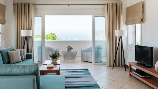 A modern living room with a sofa, a coffee table, a TV stand, and a view of the sea through large glass doors.