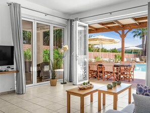 Una sala de estar moderna con televisor, muebles de madera, un comedor con asientos exteriores y vista a la piscina.